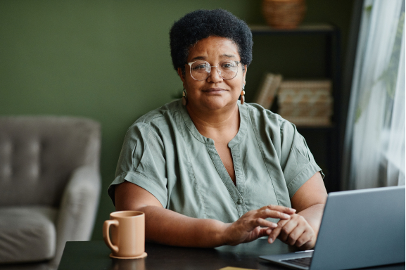 A woman sitting in a living room with a laptop in front of her, looking at you