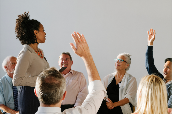 A woman moderating a community meeting. Community members are raising their hands to share their thoughts.