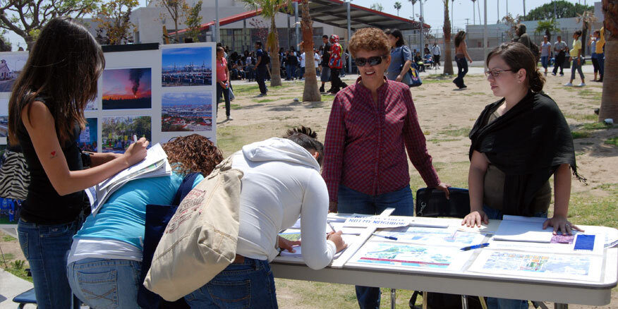 DTSC staff stand behind a table in a park. The table has displays and maps about a nearby project. 3 people are filling out forms at the table.