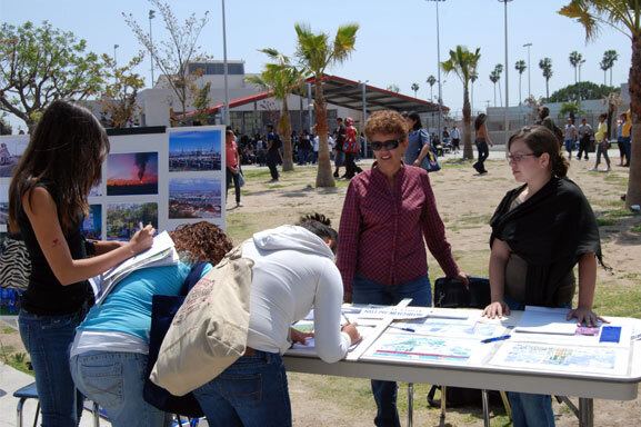 DTSC staff stand behind a table in a park. The table has displays and maps about a nearby project. 3 people are filling out forms at the table.