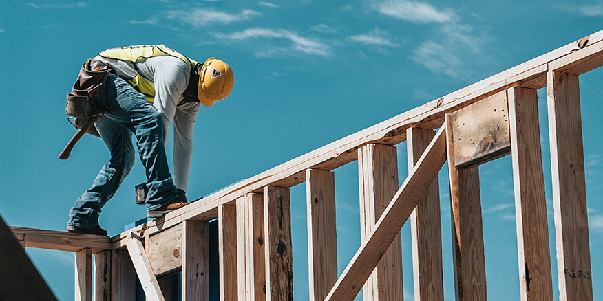 A construction worker stands on the frame of a wall using a tool