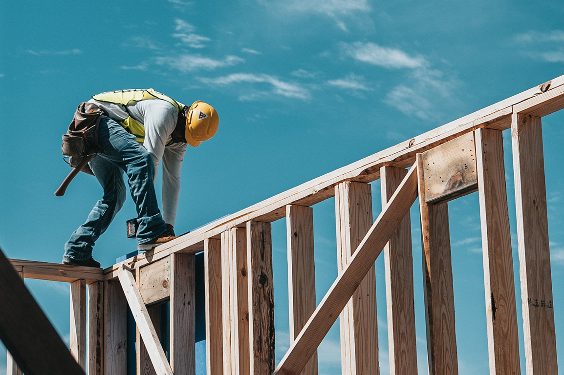 A construction worker stands on the frame of a wall using a tool