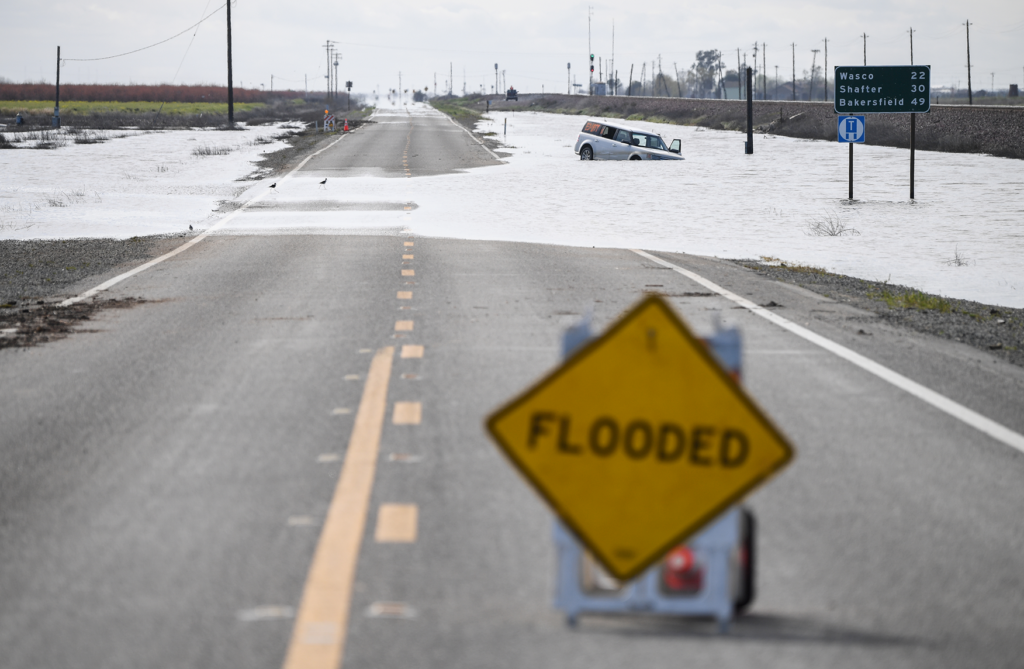 A flooded 2-lane highway in the Central Valley. A car is partially submerged on the shoulder.